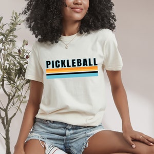 May include: A white t-shirt with a retro-style graphic that says "Pickleball" in black letters with orange and blue stripes.