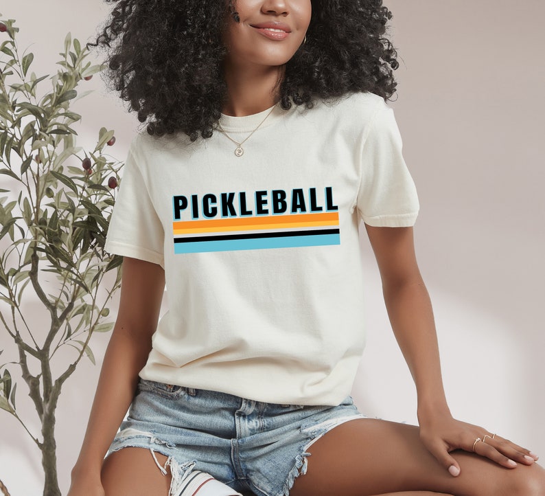 May include: A white t-shirt with a retro-style graphic that says "Pickleball" in black letters with orange and blue stripes.
