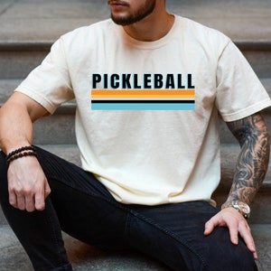 May include: A person wearing a white t-shirt with the text "PICKLEBALL" in black letters and a striped design in orange, yellow, blue, and black. The person is wearing black jeans and a bracelet on their wrist.