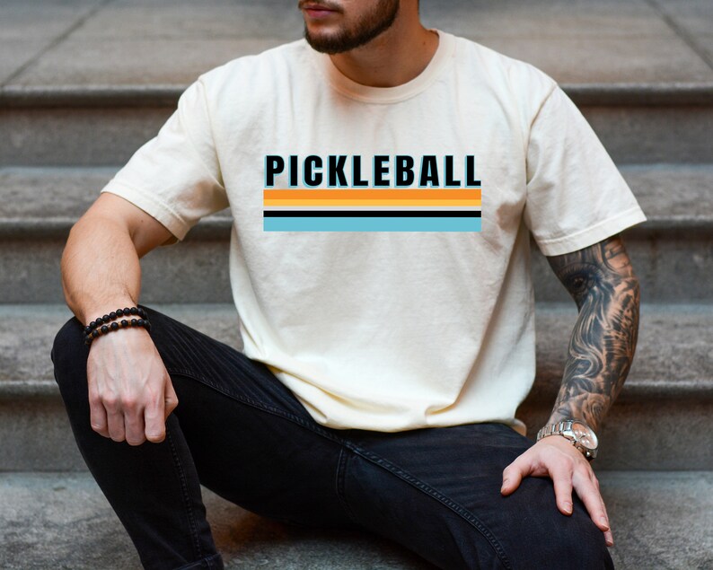 May include: A person wearing a white t-shirt with the text "PICKLEBALL" in black letters and a striped design in orange, yellow, blue, and black. The person is wearing black jeans and a bracelet on their wrist.