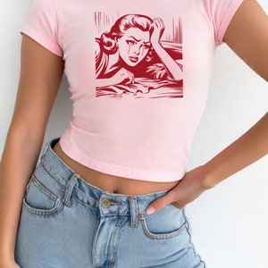 Pu&ograve; includere: Una maglietta rosa corta con una stampa grafica rossa di una donna che piange. La donna &egrave; disegnata in stile fumetto.