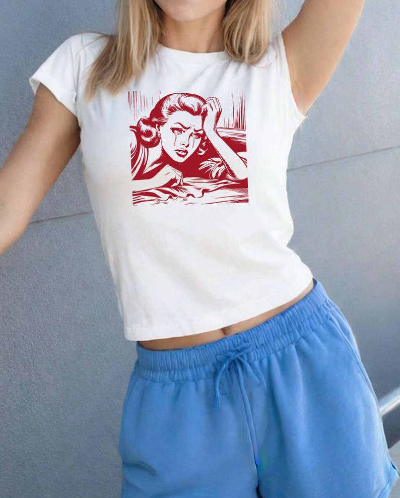 Pu&ograve; includere: Una maglietta bianca corta con una stampa grafica rossa di una donna che piange. La donna indossa un vestito rosso e ha la mano sulla fronte. La grafica &egrave; in stile fumetto.