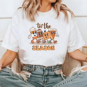 Op de afbeelding: Wit T-shirt met een retro cartoon ontwerp. De afbeelding toont een koffiekopje, een voetbal, een pompoen en een taart, allemaal samen lopend, met de tekst "Tis the Season". Het woord "SEASON" is oranje.