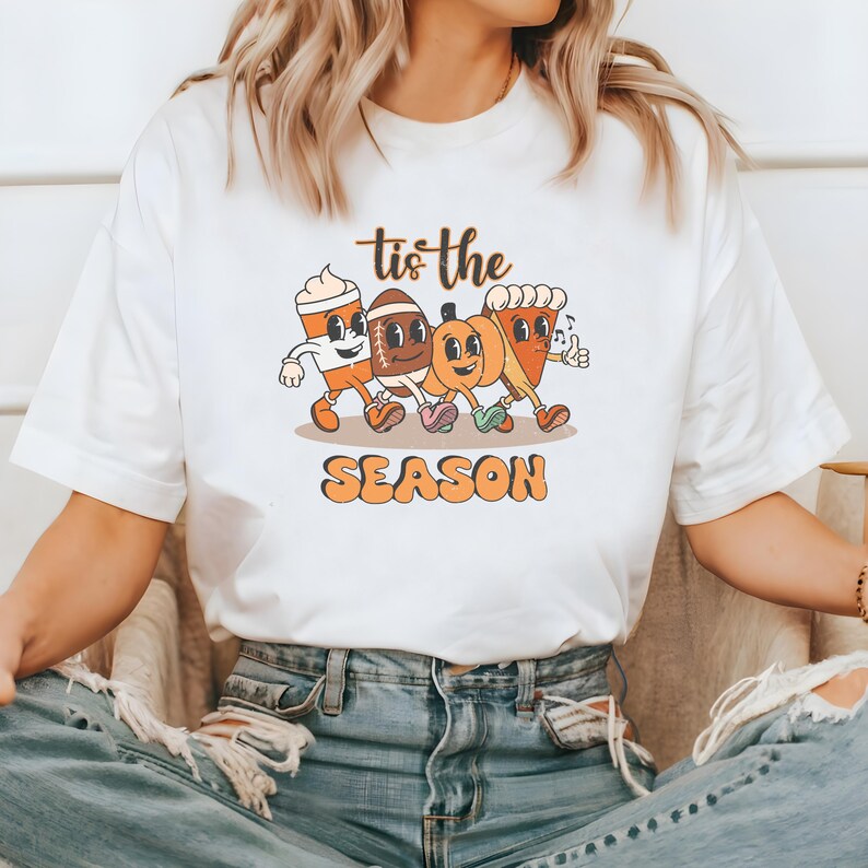 Op de afbeelding: Wit T-shirt met een retro cartoon ontwerp. De afbeelding toont een koffiekopje, een voetbal, een pompoen en een taart, allemaal samen lopend, met de tekst "Tis the Season". Het woord "SEASON" is oranje.