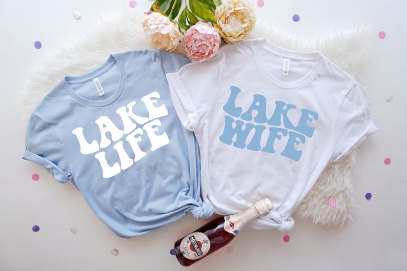 Puede incluir: Dos camisetas de color azul claro y blanco con el texto "Lake Life" y "Lake Wife" en una fuente retro. Las camisetas est&aacute;n colocadas sobre una superficie blanca y peluda con una botella de vino espumoso.