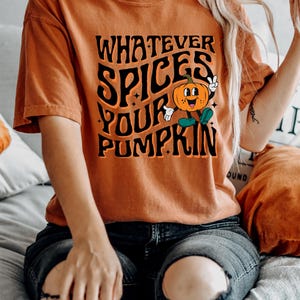May include: Orange t-shirt with the text "WHATEVER SPICES YOUR PUMPKIN" in black, featuring a cartoon pumpkin character. The shirt is a fall-themed design.