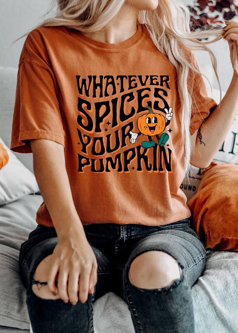 May include: Orange t-shirt with the text "WHATEVER SPICES YOUR PUMPKIN" in black, featuring a cartoon pumpkin character. The shirt is a fall-themed design.