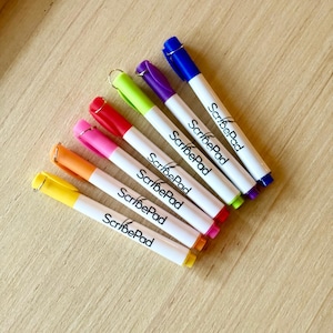 May include: A collection of seven colorful markers with white barrels, each featuring a different colored cap and tip. The text "ScribePad" is printed on the side of each marker. The markers are arranged diagonally on a light brown wooden surface.
