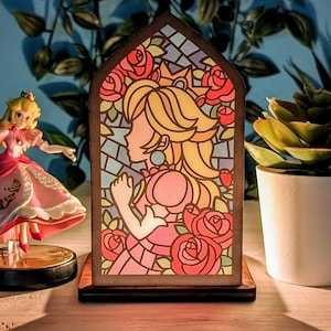 Op de afbeelding: Een lamp van gekleurd glas met een prinsesfiguur met een kroon en bloemen. De lamp is van hout en heeft een warme, uitnodigende gloed.