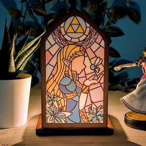 Op de afbeelding: Een lamp van gekleurd glas met een vrouwelijke figuur met blond haar en een blauwe jurk. De lamp is in een houten frame en heeft een roze, blauwe en gele kleurstelling. De lamp is aan en geeft een warm licht.