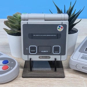 Könnte beinhalten: Eine graue und weiße Super Nintendo Entertainment System (SNES) Handheld-Konsole mit einem schwarz-weißen Ständer. Die Konsole hat einen Power-Knopf, einen Auswurftaste und einen Reset-Knopf. Die Konsole hat ein Etikett mit der Aufschrift "USE CASSETTE WITH SUPER FAMICOM MARK ONLY". Die Konsole hat ein Nintendo-Logo auf der Vorderseite.