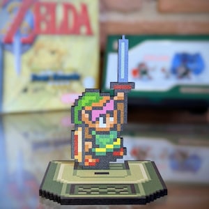Könnte beinhalten: Eine pixelartige Figur von Link aus der Videospielserie The Legend of Zelda. Link hält ein blaues Schwert und steht auf einem grün-braunen Sockel.