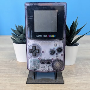 May include: A clear purple Game Boy Color handheld video game console with a black screen and the text "GAME BOY COLOR" on the front. The console is on a black stand.
