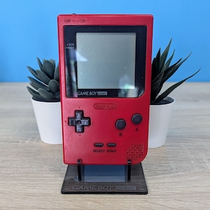 May include: A red Game Boy Pocket handheld video game console with a black and white screen. The console is on a black stand with the words "GAME BOY pocket" printed on it.