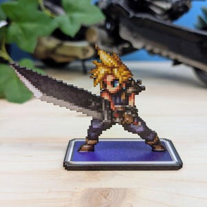 Pode incluir: Uma figura pixelizada de um personagem com cabelo loiro espetado, segurando uma grande espada preta. A figura está em uma base azul com uma borda preta. O personagem está vestindo uma roupa azul e marrom.