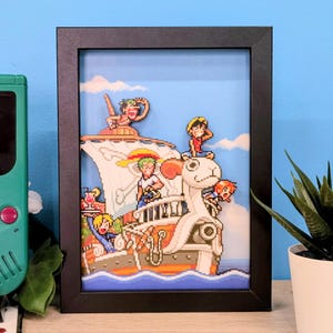 Peut inclure: Une boîte d'ombre 3D encadrée représentant des personnages pixelisés de One Piece sur le navire Going Merry. La scène inclut Luffy, Zoro, Sanji, Nami et Usopp sur un vaisseau en bois avec une figure de proue en forme de mouton sur un ciel bleu.