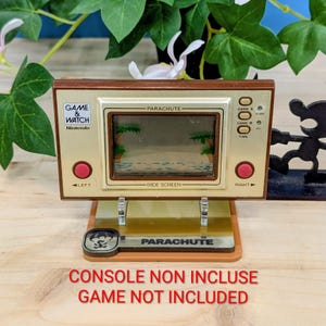 Peut inclure: Une console de jeu portable Nintendo Game & Watch "Parachute" vintage. La console dorée présente un écran affichant une scène de plage, avec des boutons rouges et le texte "GAME & WATCH" et "Nintendo". La base indique "PARACHUTE".