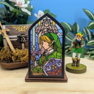 Op de afbeelding: Houten decoratie in glas-in-loodstijl met een personage uit The Legend of Zelda, met een zwaard en schild. Het ontwerp maakt gebruik van groene, blauwe en paarse tinten. Een klein beeldje van hetzelfde personage is ook zichtbaar, samen met een schatkist.