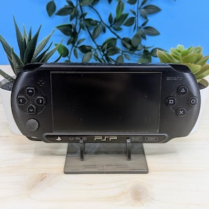 Peut inclure: Une console de jeu portable Sony PSP noire sur un support en acrylique transparent. La console a un écran noir et des boutons, et le support a le mot "PSP" imprimé dessus.