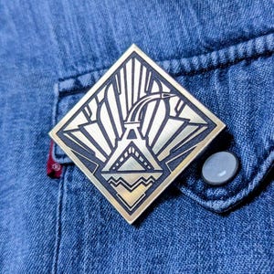 Puede incluir: Un pin dorado en forma de diamante con un borde negro y un diseño art déco. El pin está sujeto a una chaqueta vaquera azul, cerca del bolsillo y un botón gris.