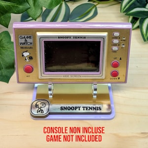 Puede incluir: Consola portátil vintage Snoopy Tennis Game & Watch, con marco dorado y rosa y botones rojos. El juego se muestra en un soporte con el logotipo de Snoopy Tennis. El texto "CONSOLE NON INCLUSE GAME NOT INCLUDED" es visible.