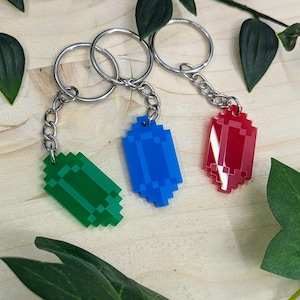 May include: Three keychains with pixelated gemstone charms in green, blue, and red. Each charm is attached to a silver chain and ring. The keychains are displayed on a light wood surface with green leaves.