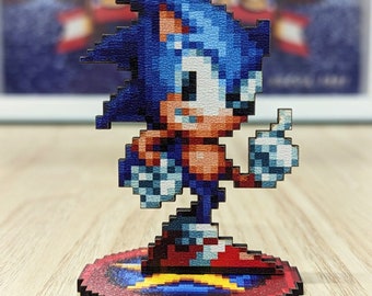 May include: A blue and red pixelated figurine of Sonic the Hedgehog standing on a circular base with a star design. The figurine is made of wood and has a 3D effect.