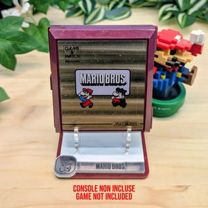 Może przedstawiać: Bordowa konsola Nintendo Game & Watch z logo i postaciami Mario Bros. Konsola jest wyświetlana na przezroczystym stojaku, z figurką Mario w tle. Tekst "CONSOLE NON INCLUSE GAME NOT INCLUDED" znajduje się na dole.