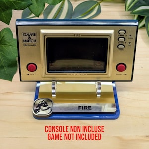 Puede incluir: Una consola portátil Game & Watch dorada y azul con la palabra "FIRE" en el soporte. La consola tiene una pantalla negra, botones rojos y el texto "GAME & WATCH Nintendo". El soporte tiene una base azul y un emblema plateado.
