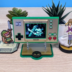 Puede incluir: Una consola de videojuegos portátil Game & Watch: The Legend of Zelda verde y dorada con un soporte. La consola presenta una pantalla que muestra a Link balanceándose en una liana. La consola está en un soporte verde y dorado con un logotipo de Triforce.