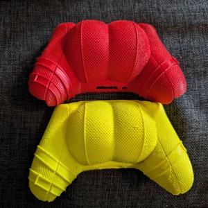 Może przedstawiać: Dwa kolorowe uchwyty do kontrolera, jeden w kolorze jasnoczerwonym, drugi żółtym. Oba mają teksturowaną powierzchnię i są przeznaczone do kontrolera do gier. Leżą na ciemnoszarej tkaninie.