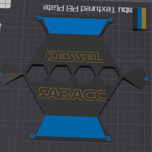 Puede incluir: Una placa PEI negra y geométrica con las palabras "KESSEL" y "SABACC" en letras doradas. La placa tiene detalles azules y está sobre un fondo de rejilla gris oscuro. Las palabras "nbu Textured PEI Plate" son visibles.