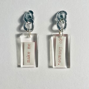 May include: A pair of clear rectangular earrings with the words "make me" and "go insane." printed in red. The earrings have silver chain links at the top.