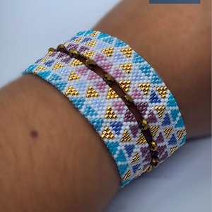 May include: Two beaded bracelets with a geometric pattern in shades of blue, white, pink, and gold. The bracelets are stacked on a person's wrist.