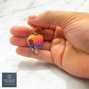 Puede incluir: Un pendiente hecho a mano con cuentas en forma de lágrima, sostenido en una mano. El pendiente presenta un degradado de colores, incluyendo amarillo, naranja, rosa y azul. El pendiente está hecho con pequeñas cuentas y tiene un gancho dorado.