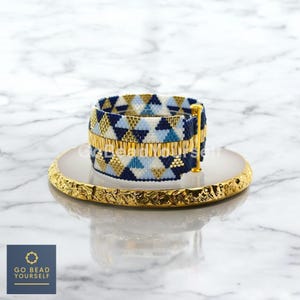 Puede incluir: Un brazalete de cuentas con un patrón geométrico de triángulos azules, blancos y dorados. El brazalete está sobre una superficie de mármol blanco con un borde dorado.