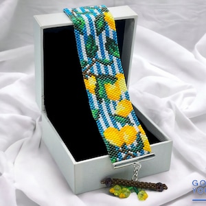 Könnte beinhalten: Ein Armband aus Perlen mit einem Zitronen- und Blattdesign in den Farben Gelb, Grün, Blau und Weiß. Das Armband befindet sich in einer weißen Schachtel mit schwarzem Innenraum. Das Armband hat einen silbernen Verschluss und einen Perlenanhänger.