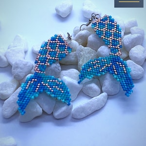 May include: A pair of blue and pink mermaid tail earrings with silver hooks. The earrings are made of beads and are displayed on a white background.
