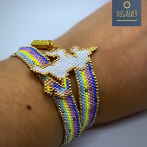 Puede incluir: Un brazalete de cuentas de arcoíris con un dije de unicornio blanco. El brazalete está hecho con cuentas pequeñas y tiene un cierre dorado. El dije está unido al brazalete con un anillo dorado. El brazalete está en la muñeca de una persona.