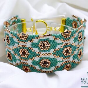May include: A beaded bracelet with a geometric pattern in teal, white, and copper colours. The bracelet features a gold-coloured clasp and decorative copper-coloured accents. The bracelet is displayed on a white surface.