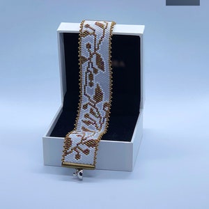 Peut inclure: Un bracelet blanc en perles avec un motif floral doré et brun. Le bracelet est dans une boîte blanche avec un intérieur noir. La boîte porte une étiquette qui dit "GO BEAD YOURSELF".