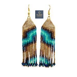 Puede incluir: Pendientes de flecos de cuentas doradas y azules con un patrón geométrico. Los pendientes miden aproximadamente 7,5 cm de largo.