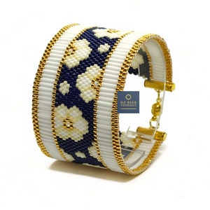 Peut inclure: Bracelet en perles blanches et bleu marine avec des accents dorés et un motif floral. Le bracelet est sur un présentoir transparent. "GO BEAD YOURSELF" est imprimé sur un cercle blanc.