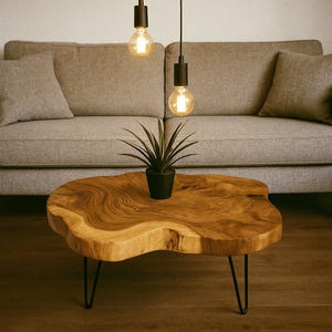 Pode incluir: Uma mesa de centro de madeira com borda natural, padrão de veios e bordas arredondadas irregulares, apoiada por três pernas finas de metal preto em estilo hairpin. Um pequeno vaso preto com uma planta verde pontiaguda está no centro.