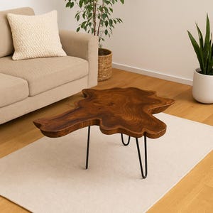 Pode incluir: Uma mesa de centro única com uma superfície de madeira castanha escura de borda natural e pernas de hairpin pretas. A mesa tem uma forma orgânica e irregular e está sobre um tapete de cor clara. A mesa está perto de um sofá bege e uma planta de interior.
