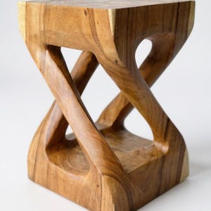 May include: A handcrafted wooden stool with a unique, intertwined design. The natural wood grain is visible, showcasing the warm brown tones of the material. The stool's dimensions are approximately 12 inches square and 18 inches tall.