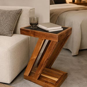 Op de afbeelding: Een houten bijzettafel met een uniek Z-vormig ontwerp. De tafel heeft een donker blad, een glas en twee boeken. De warme houttinten contrasteren met de neutrale achtergrond en creëren een moderne esthetiek. De tafel is een stijlvolle toevoeging aan elke woonruimte.