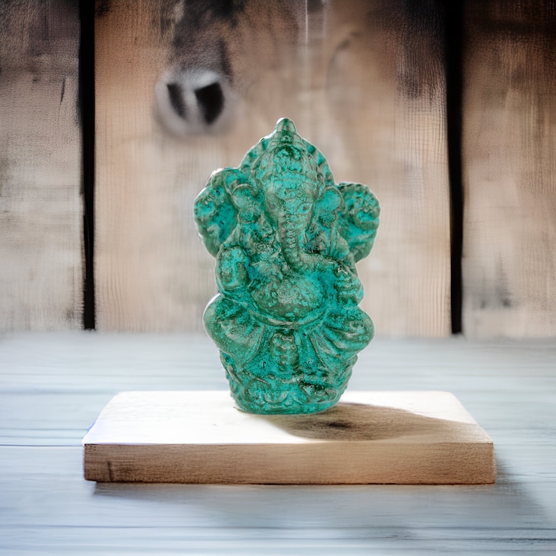 May include: A turquoise-colored statue of the Hindu deity Ganesha, seated in a meditative pose. The statue has intricate details and is placed on a small, rectangular wooden block. The background is a blurred wooden surface.