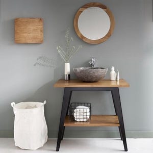 Può includere: Un mobile da bagno con piano in legno e gambe in metallo nero. Il mobile ha un lavabo rotondo in pietra, uno specchio rotondo con cornice in legno e un cesto per la biancheria in tela bianca.