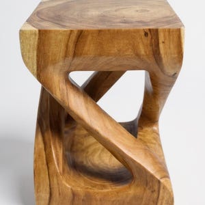 May include: A solid wood stool with a unique, sculptural design. The square seat rests on a base with a carved, openwork pattern. The natural wood grain and warm tones create a rustic, yet modern aesthetic. This piece is suitable for seating or as a decorative accent.